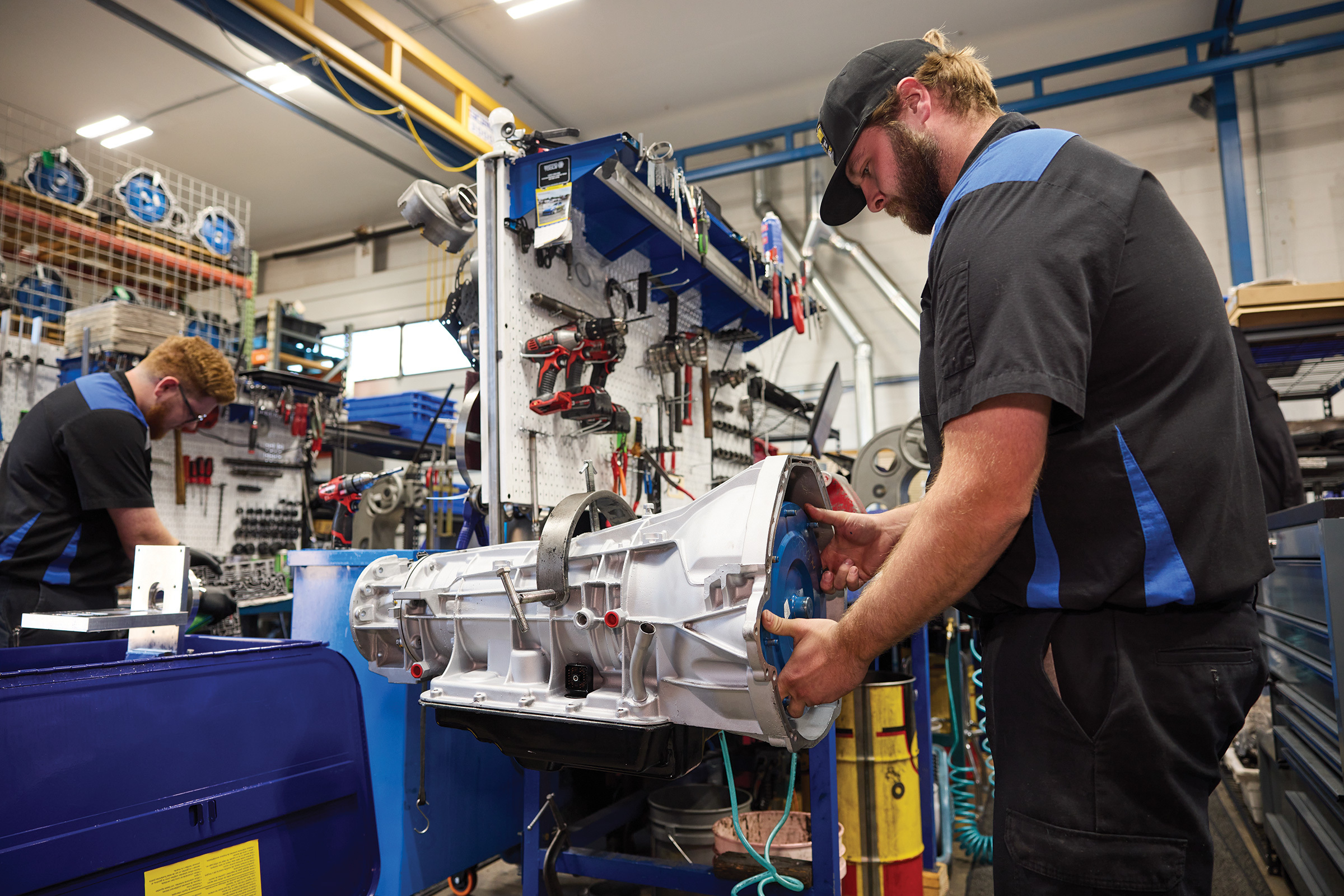 Mechanic in black and blue uniform installing a torque converter into a transmission in a busy workshop.