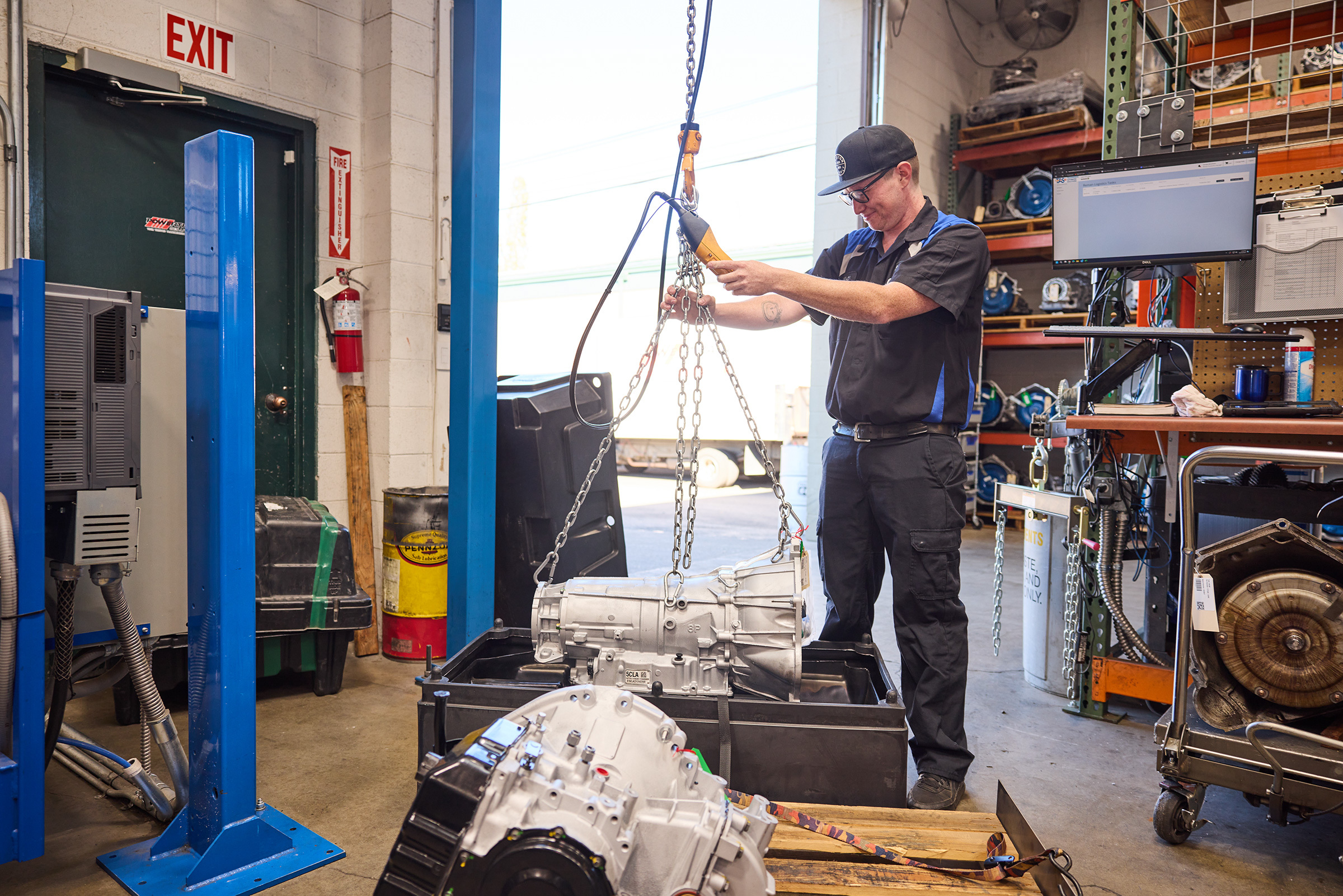 Auto technician using a hoist to lift a transmission unit in a well-organized workshop.