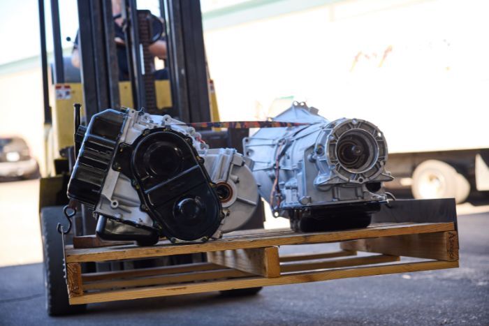A forklift transports two automotive transmissions on a wooden pallet in an outdoor industrial setting.
