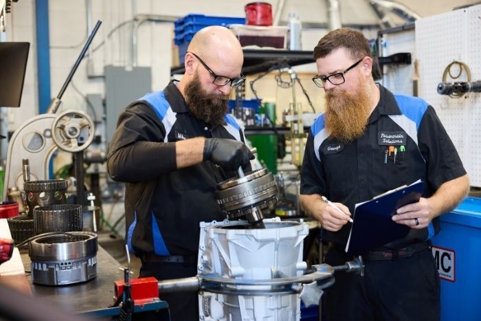 Two technicians examine and assemble machinery parts at a workbench in a well-equipped repair shop.