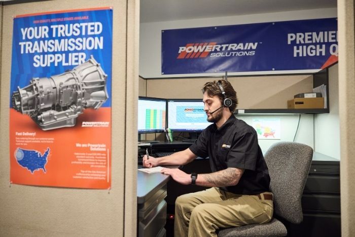 Office worker at a desk with computer screens, surrounded by Powertrain Solutions and transmission supplier posters.