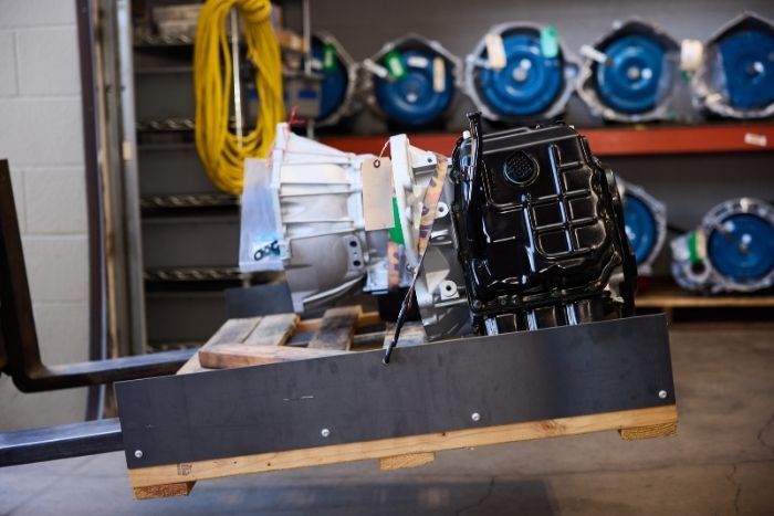 Automotive powertrain assembly on a pallet with transmission units on shelves in the background.